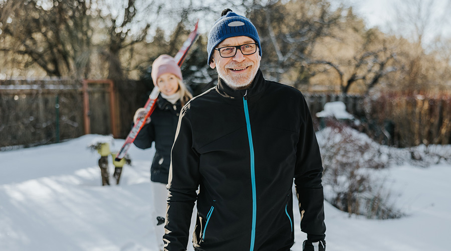 A man in a winter coat standing in the snow with a woman holding skis standing behind him.