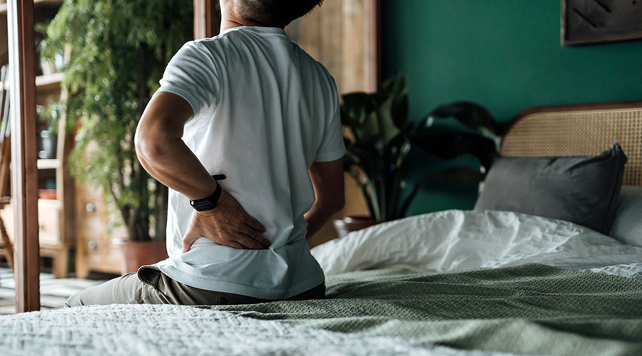 Man sitting on the edge of the bed, holding his lower back in pain and discomfort.